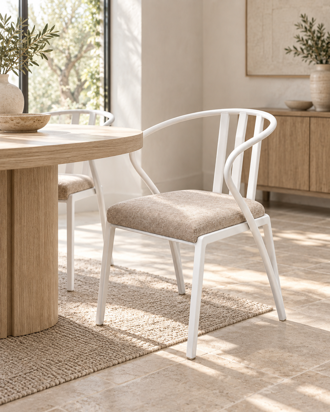 White metal armchair handcrafted in Kenya, with curved spindle back and oatmeal cushioned seat, beside a round timber table on stone flooring